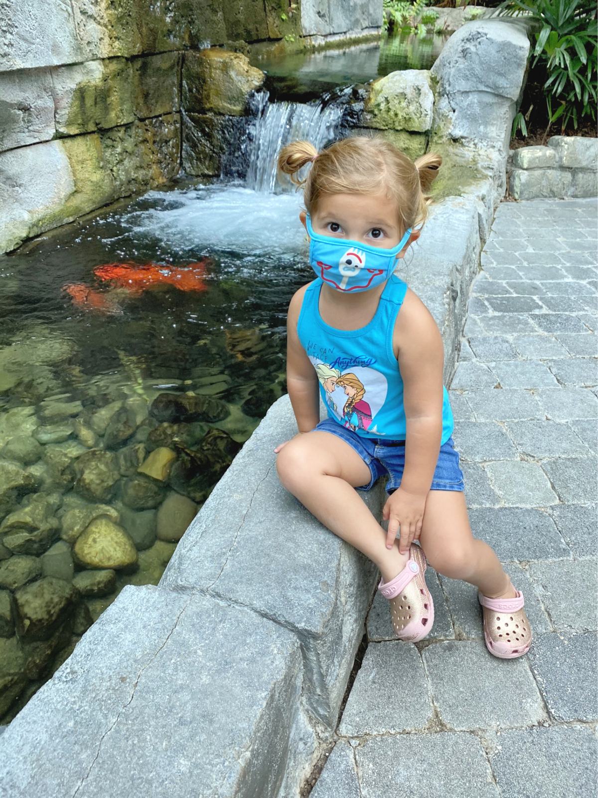 Little girl with blue mask on face sitting next to a koi pond.