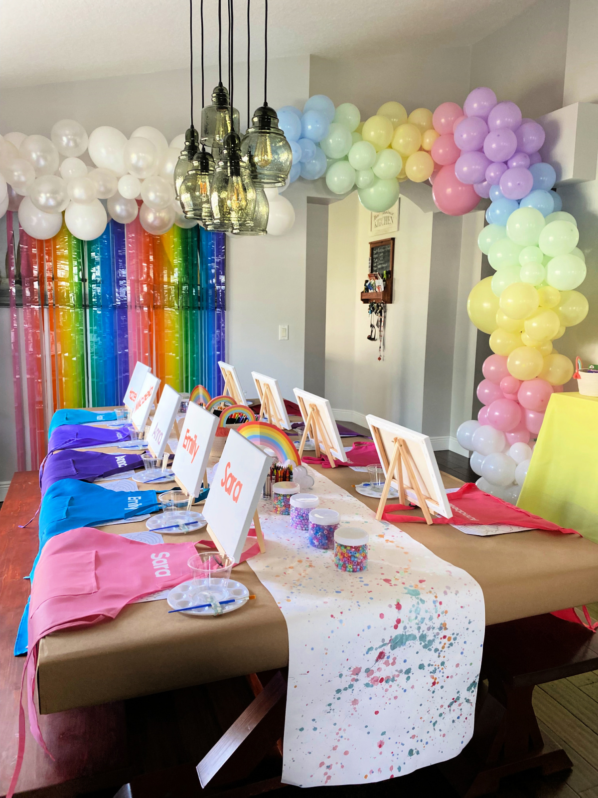 Pastel rainbow balloon arch with long brown table with aprons, easels, canvases, beads, and placemats.