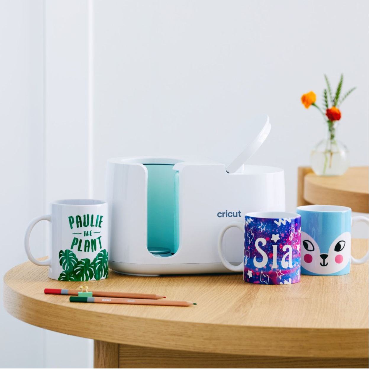 Cricut mug press with three custom mugs on a light wood table.