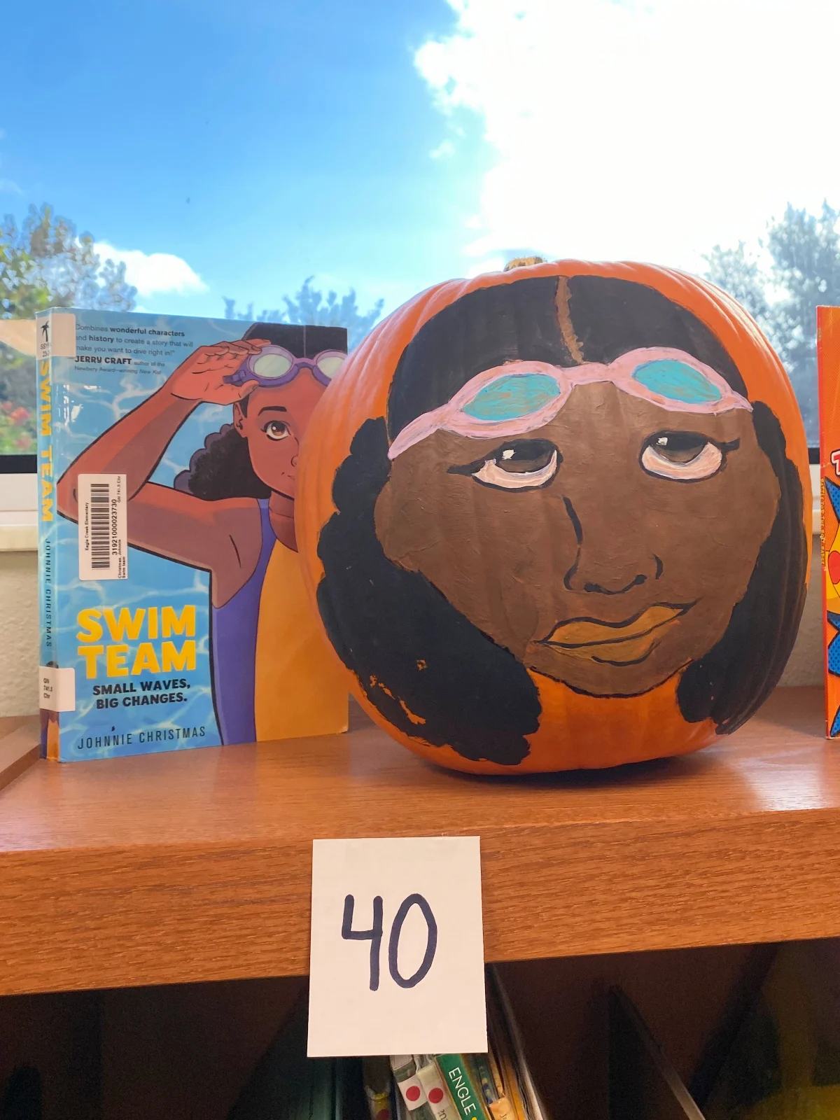 Orange pumpkin with an african american face on the pumpkin next to a book called "swim team"