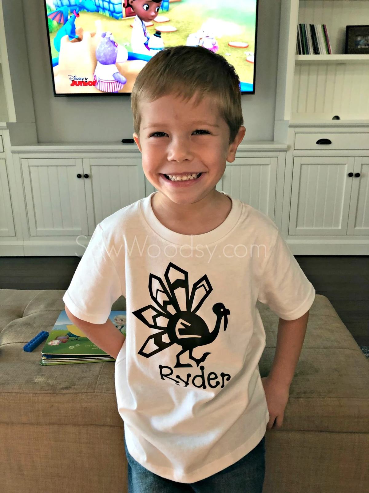 Little boy with a turkey shirt leaning up against a tan ottoman.