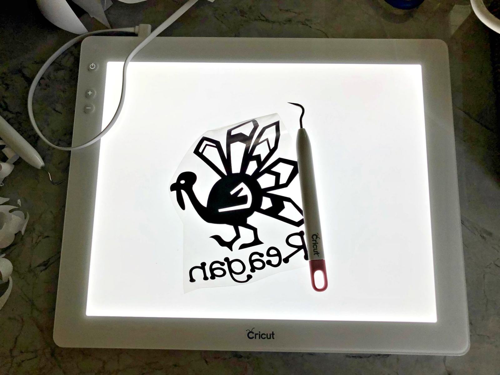 Cricut BrightPad with iron on vinyl cut out and weeding tool on marble counter.