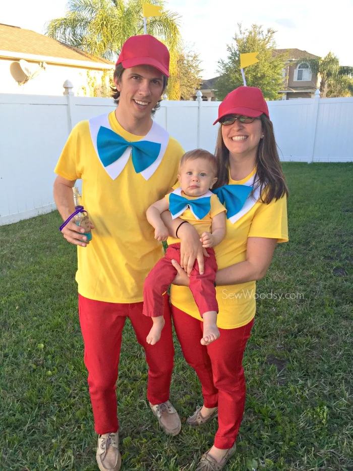 Male, female, and baby dressed as Tweedledum and Tweedledee standing in a green yard with white fence.