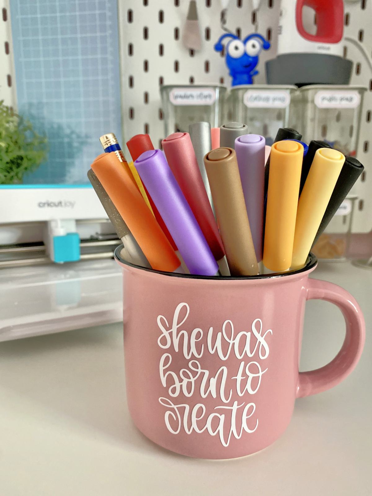 Pink mug that says "she was born to create" with pens in mug.