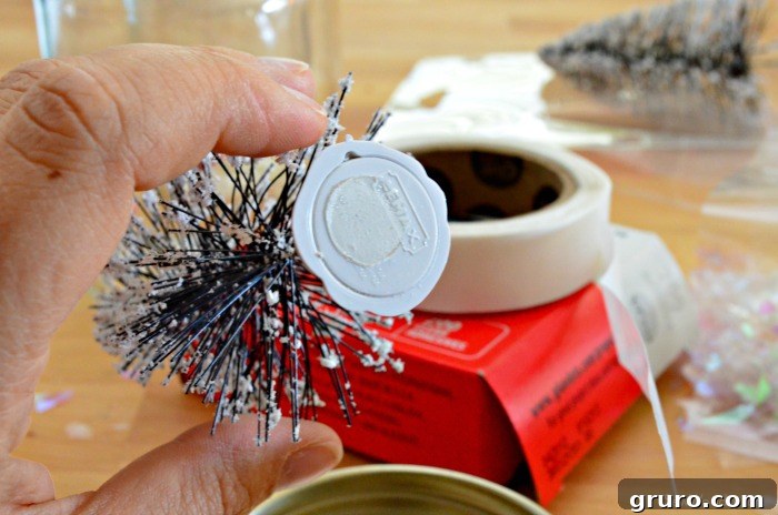 A female hand holds a miniature wire bottle brush tree, carefully applying a small, clear glue dot to the very center of its base for secure adhesion.