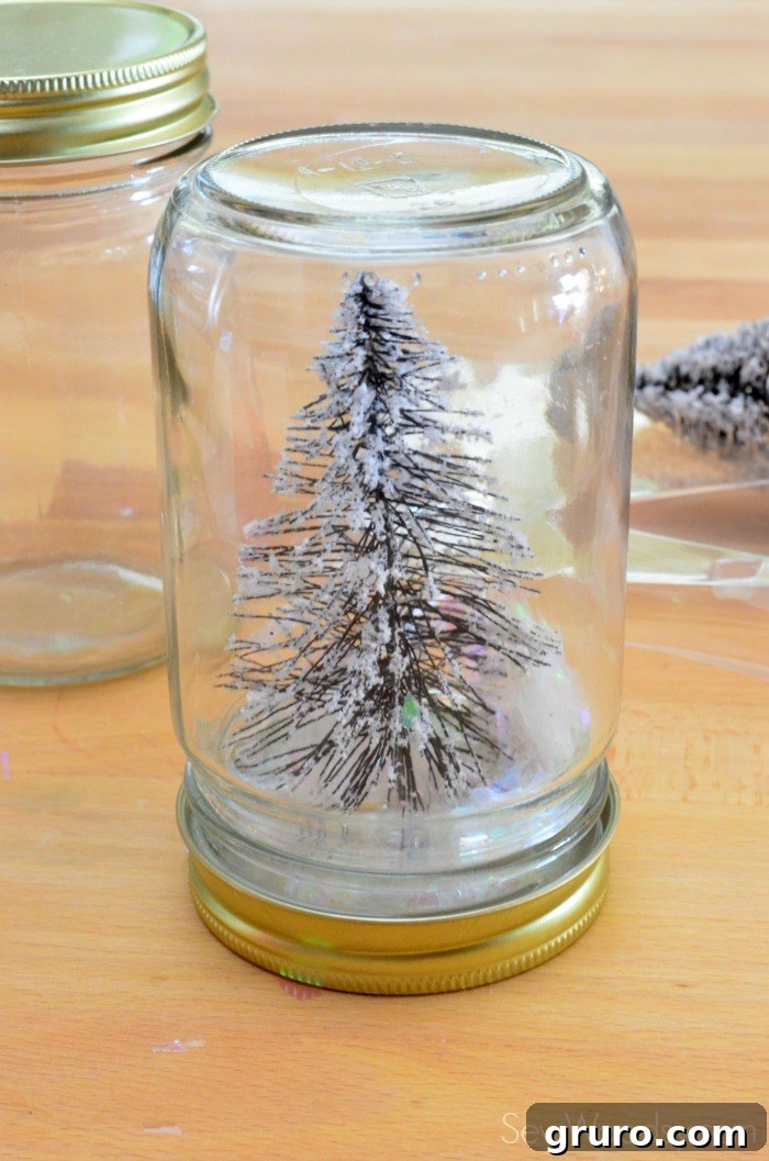 A mason jar lid, with a miniature Christmas tree attached inside, is being carefully screwed onto the glass jar filled with artificial snow.