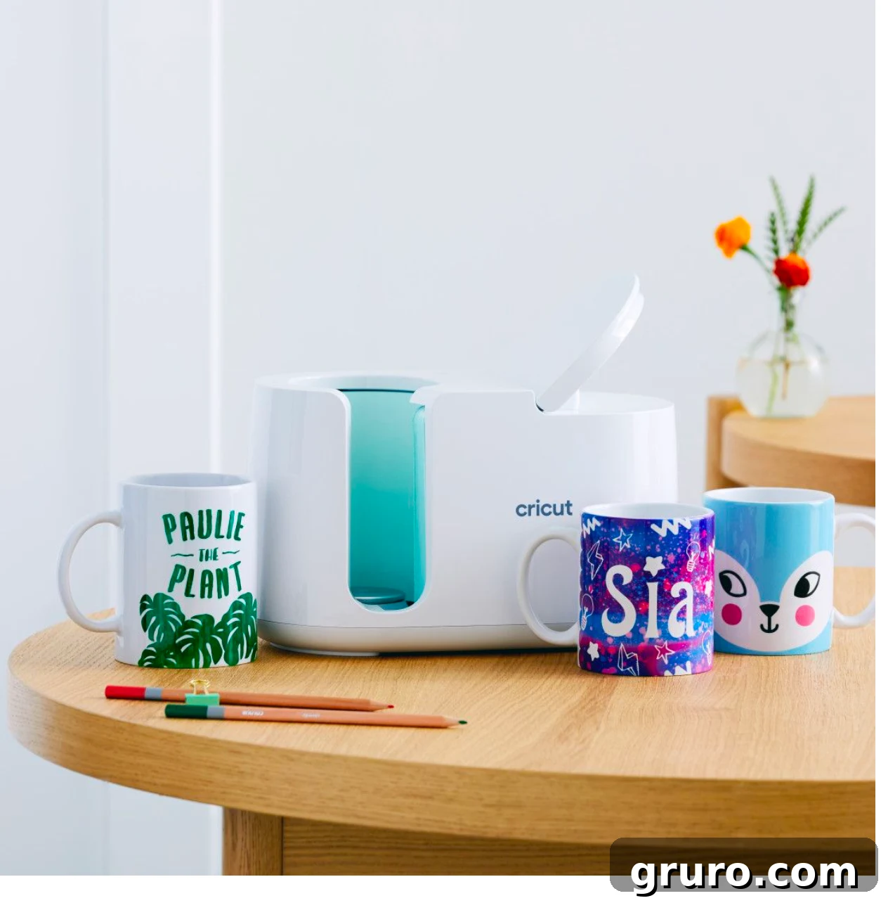 A sleek Cricut Mug Press, shown on a light wood table alongside three beautifully customized mugs, demonstrating its ease of use and professional results.
