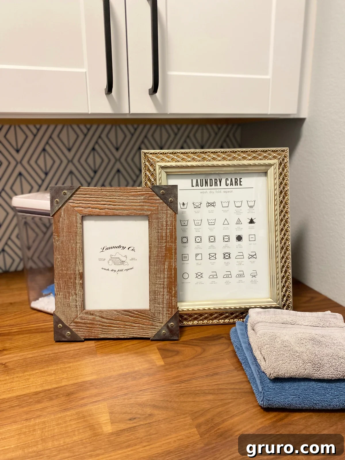 Your Printable Laundry Cheat Sheet 2 Two framed printables, one with laundry symbols and one saying 'Laundry Co.', sitting on a wooden countertop with neatly stacked white towels.