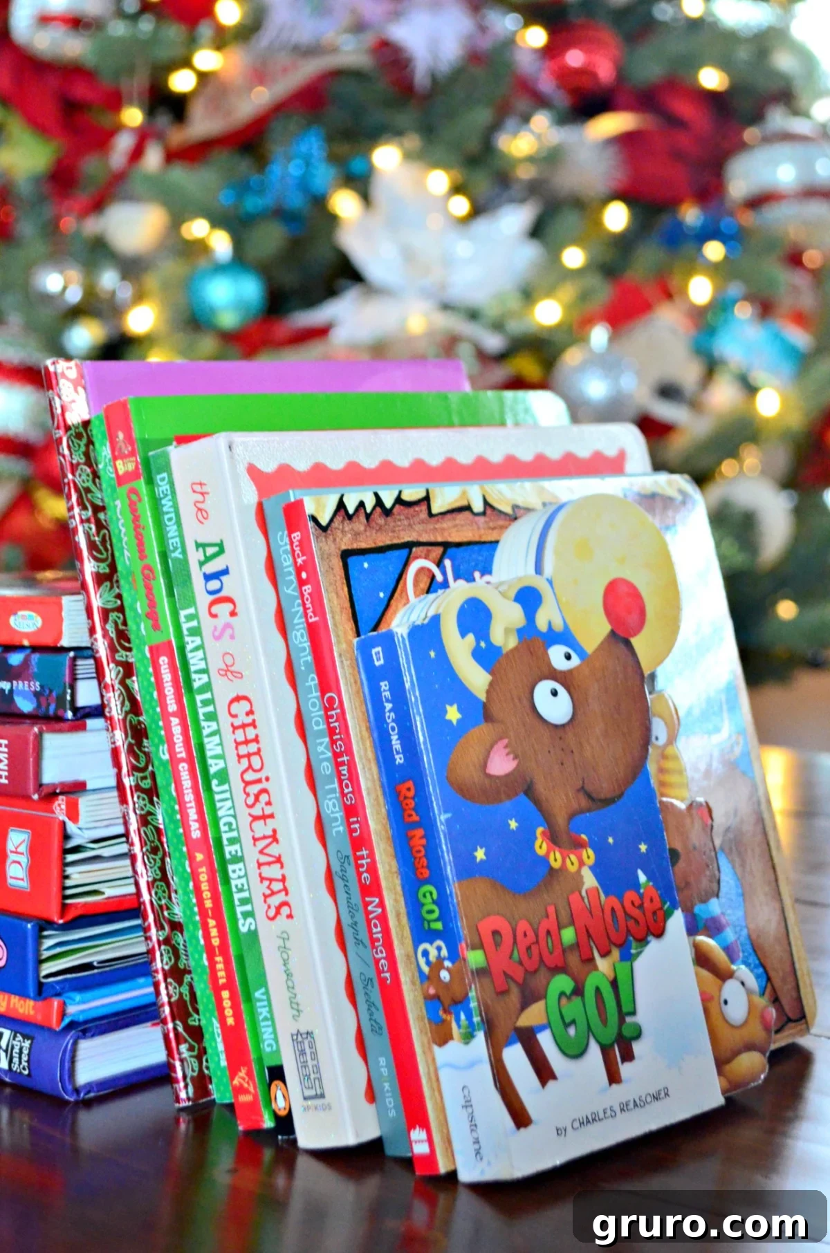 A stack of Christmas books on a wood table with a christmas tree in the background.