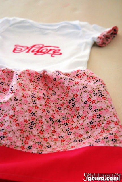 White onesie with the word "adore" on it, featuring a charming pink floral fabric skirt and delicate trim.