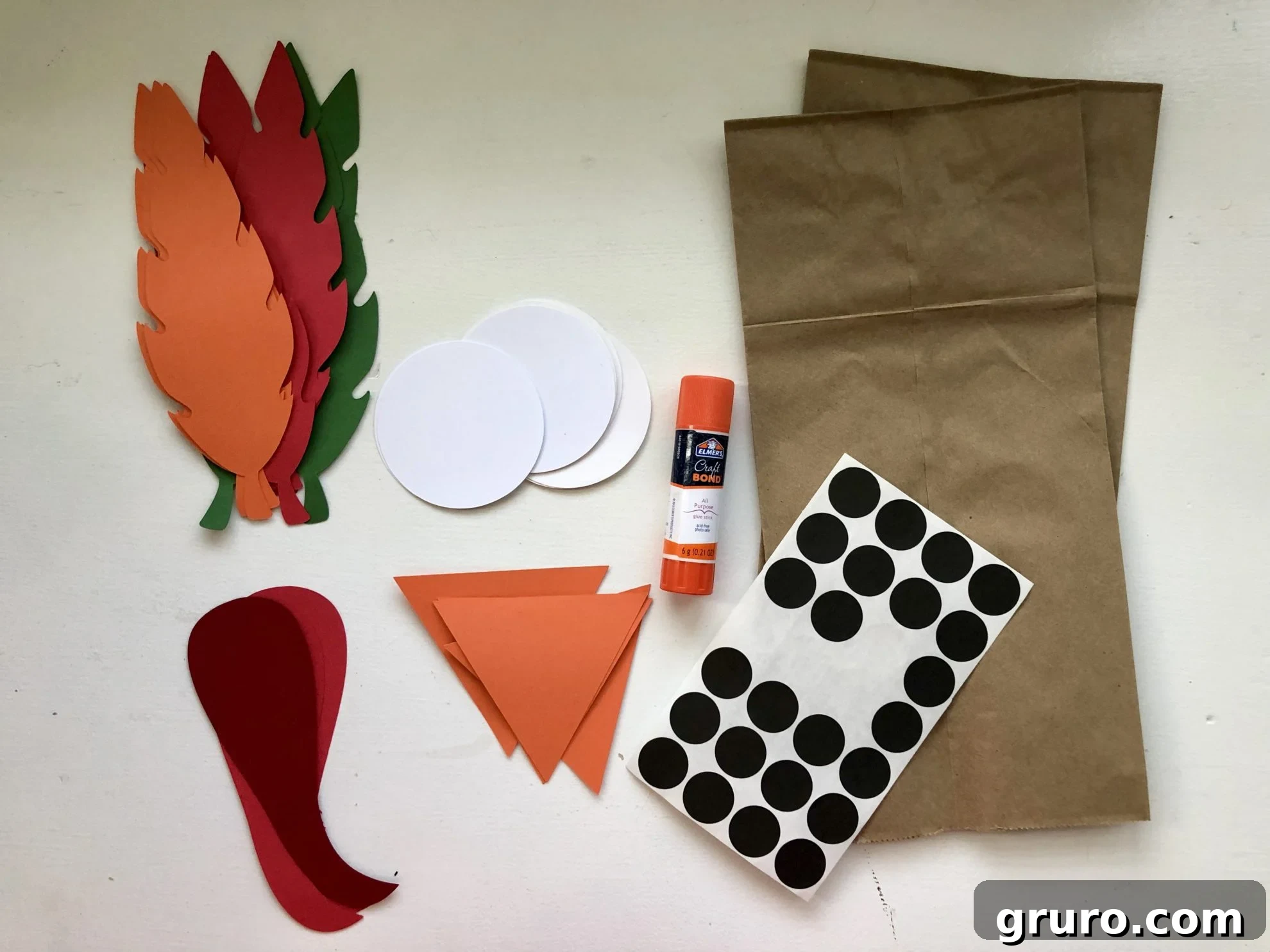 DIY Turkey Paper Bag Puppets 3 An organized display of craft supplies, including colorful paper feathers, white circles for eyes, orange triangles for beaks, red gobblers, a glue stick, black sticker dots, and sturdy brown paper bags, all ready for assembly.