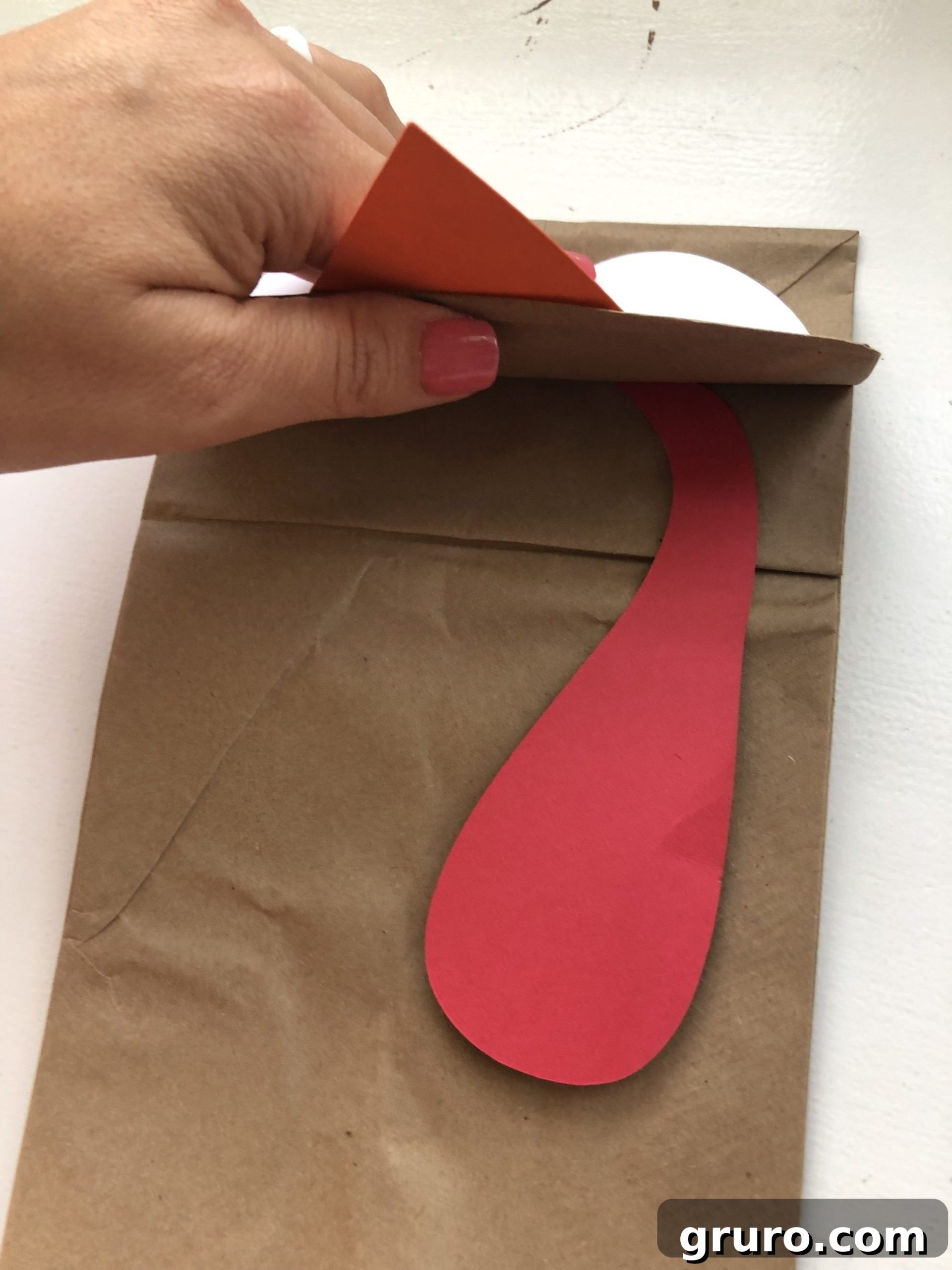DIY Turkey Paper Bag Puppets 7 A gentle female hand with pink nail polish carefully tucking a red paper gobbler beneath the folded flap of a brown paper bag, giving the turkey its characteristic wattle and a pop of color.