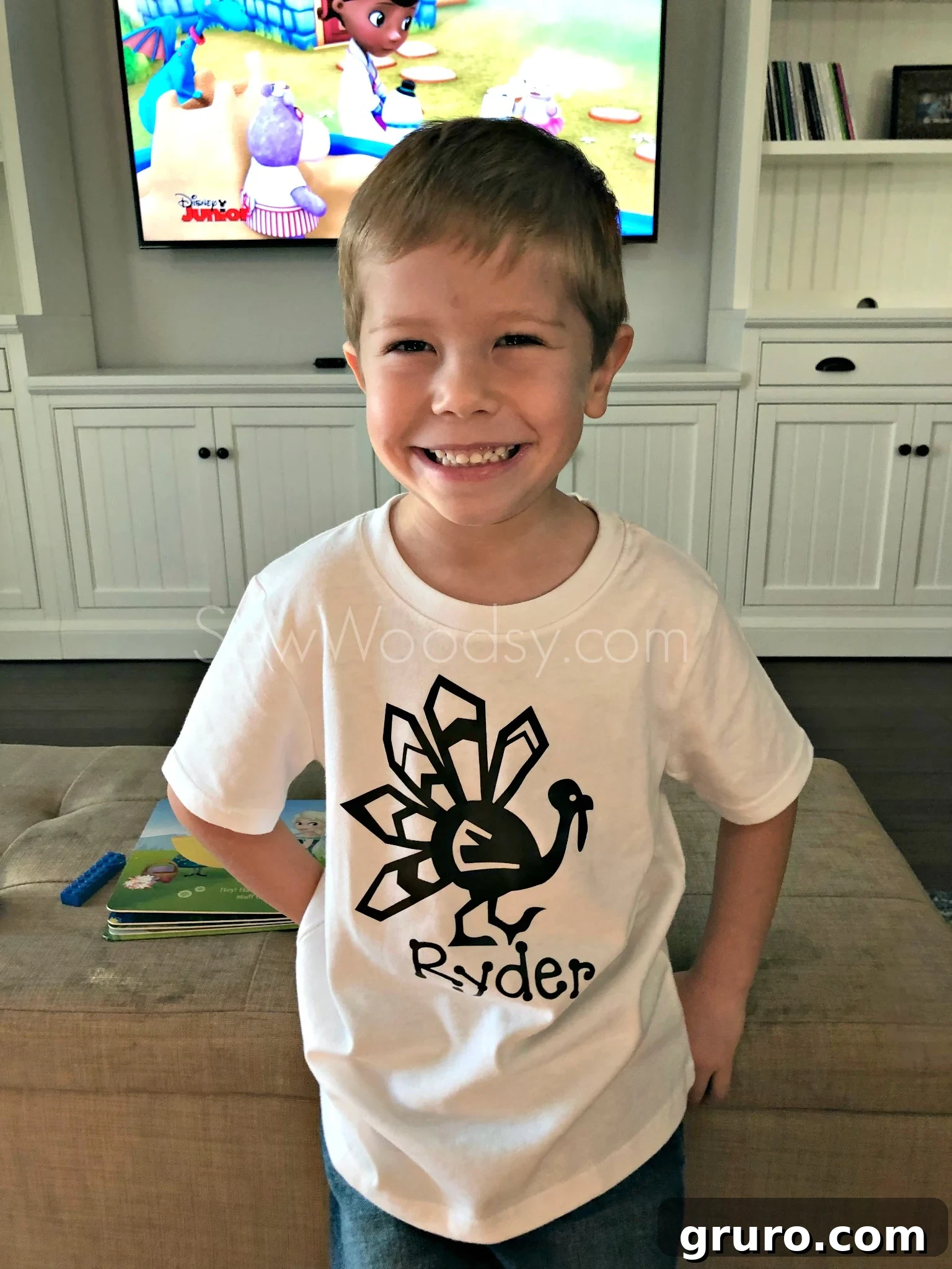 A little boy proudly wearing his personalized Thanksgiving turkey shirt, leaning against a tan ottoman, captured in a delightful portrait.