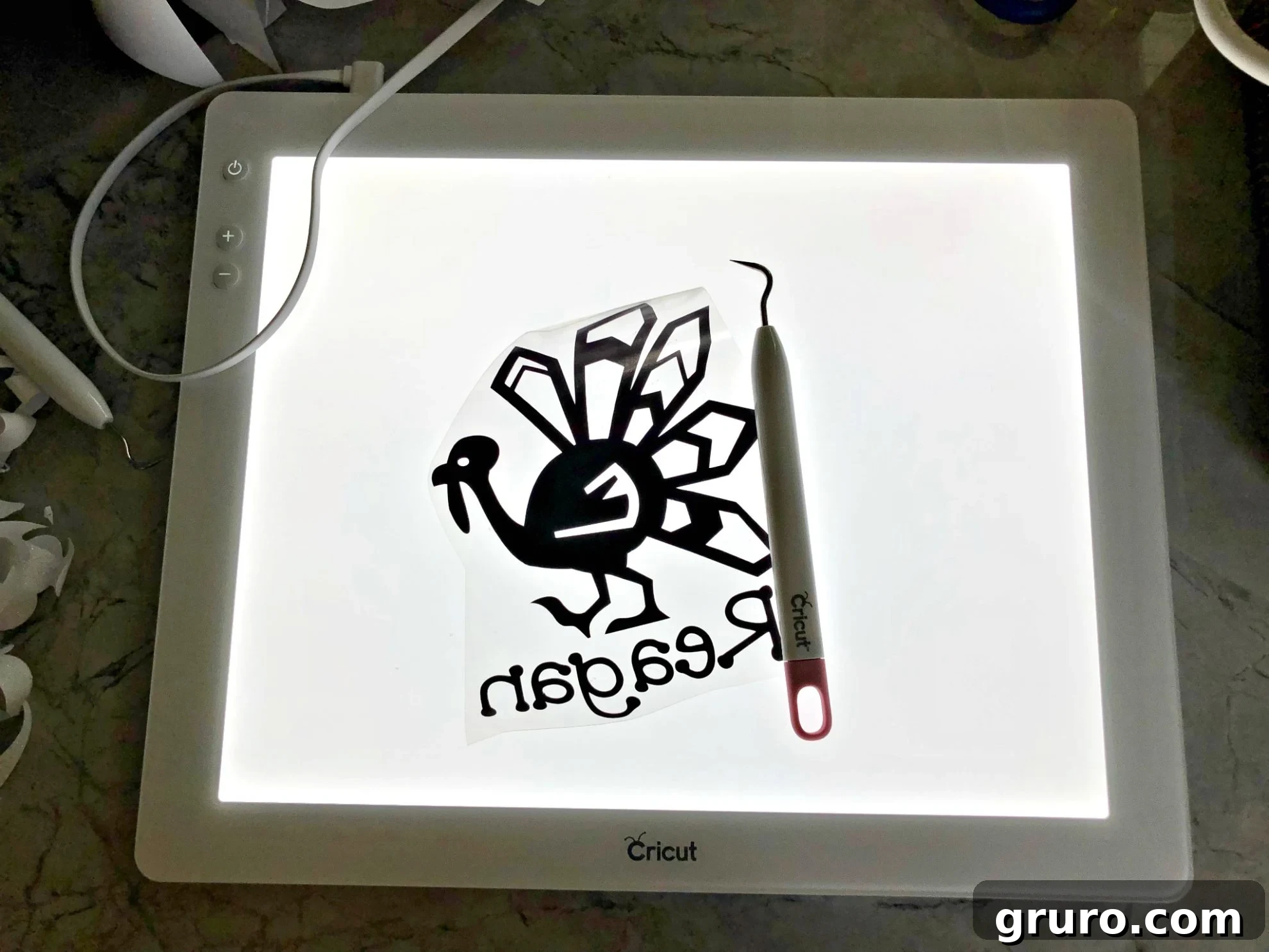 Cricut BrightPad illuminating a cut iron-on vinyl design on a marble counter, with a weeding tool poised to remove excess material.
