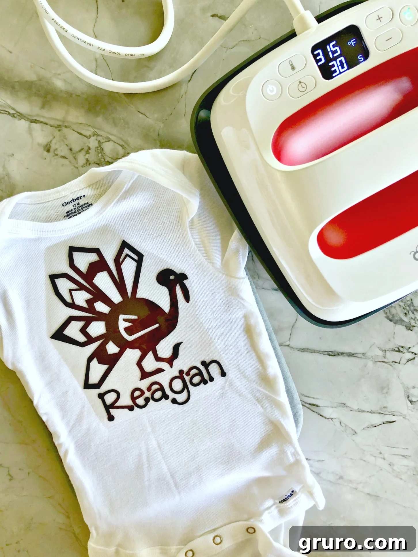 Top-down view of a white baby onesie with a personalized turkey design laid out next to a Cricut EasyPress 2, ready for heat application.