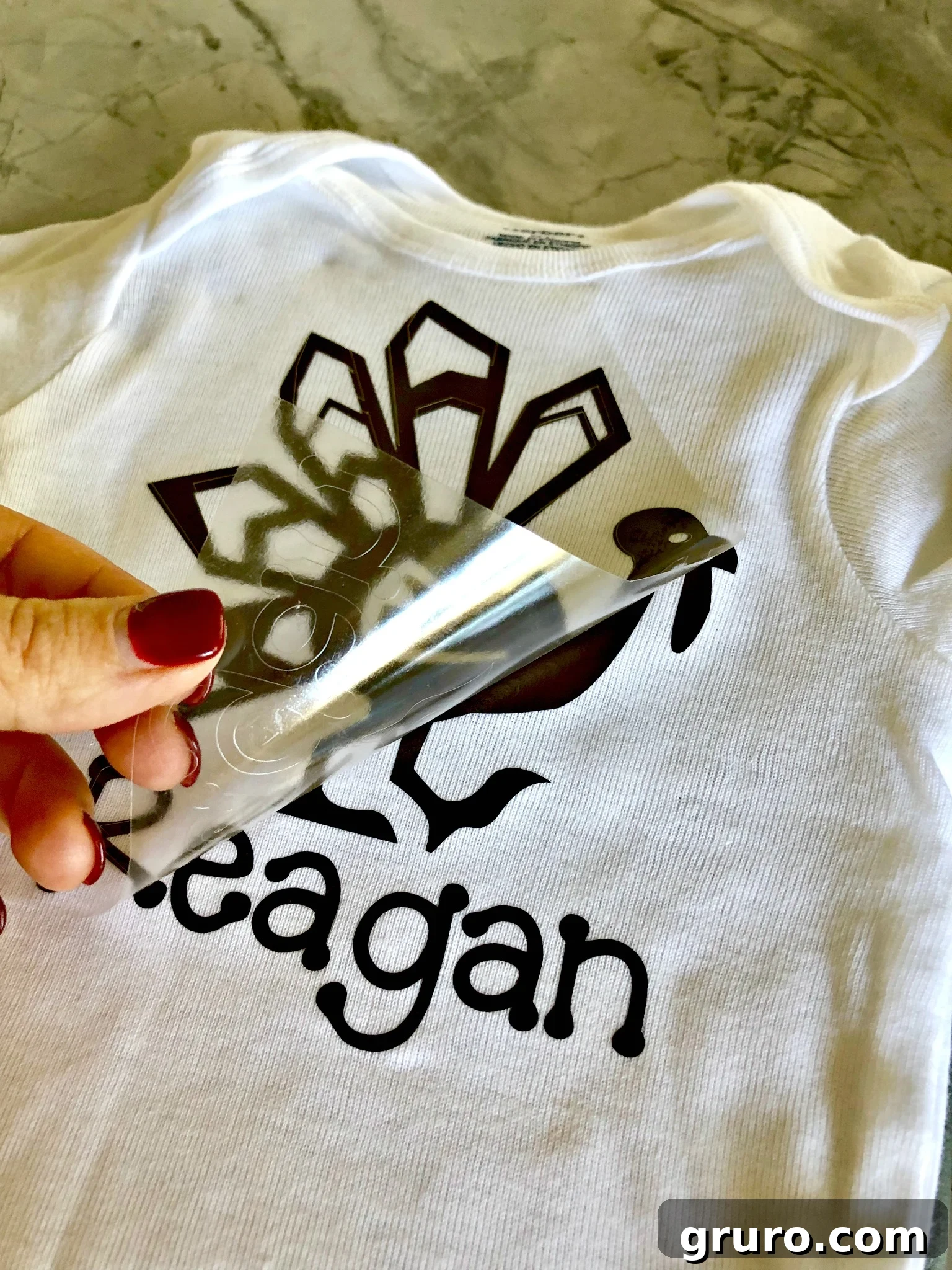 A hand with maroon nails carefully peeling off the clear plastic carrier sheet from an iron-on vinyl design on a white t-shirt, revealing the finished turkey and name.