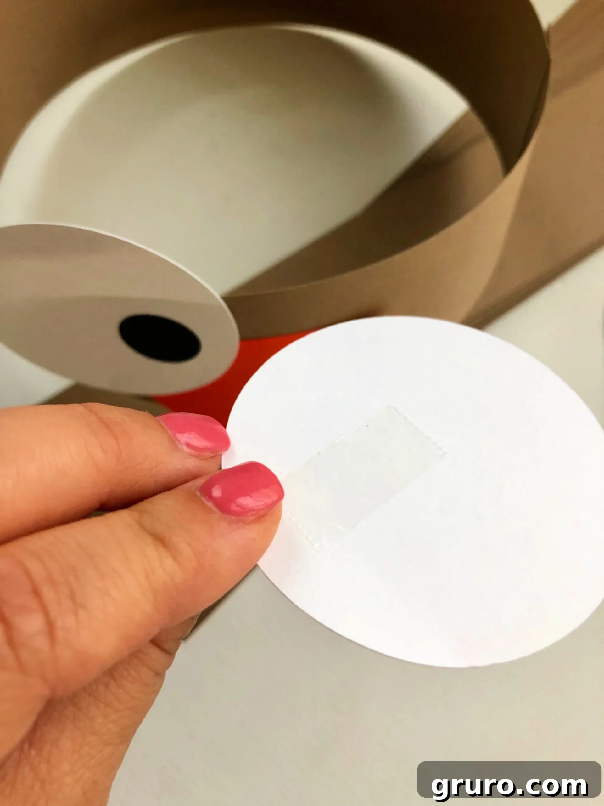 Female hand with pink nails holding a white circle with double stick tape on it.