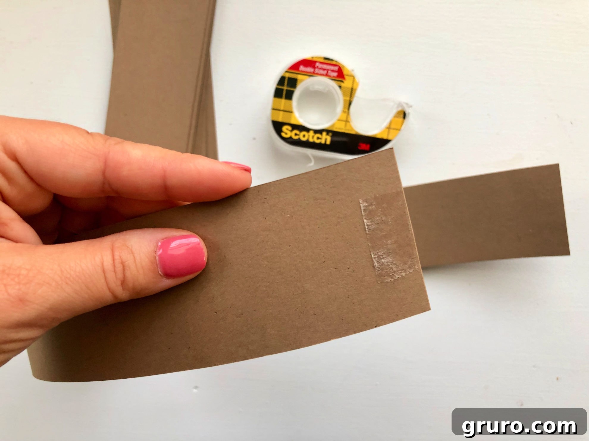 Female hand with pink fingernails holding brown cardstock strip with double sided tape on it.