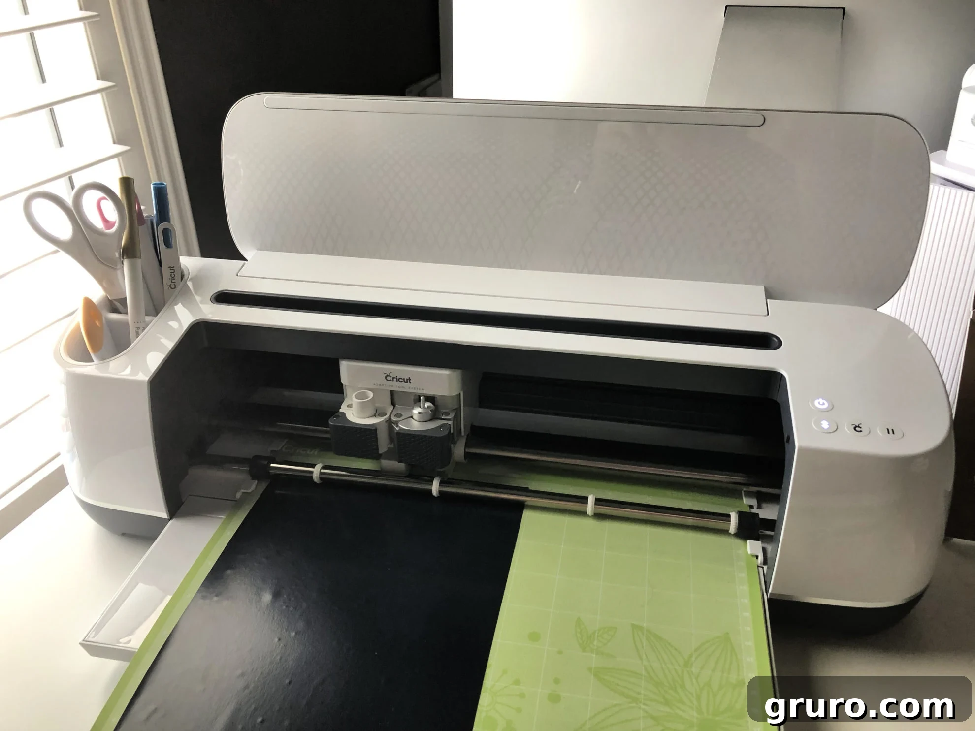 A pristine white Cricut Maker machine with a vibrant green cutting mat loaded, ready for a crafting project.