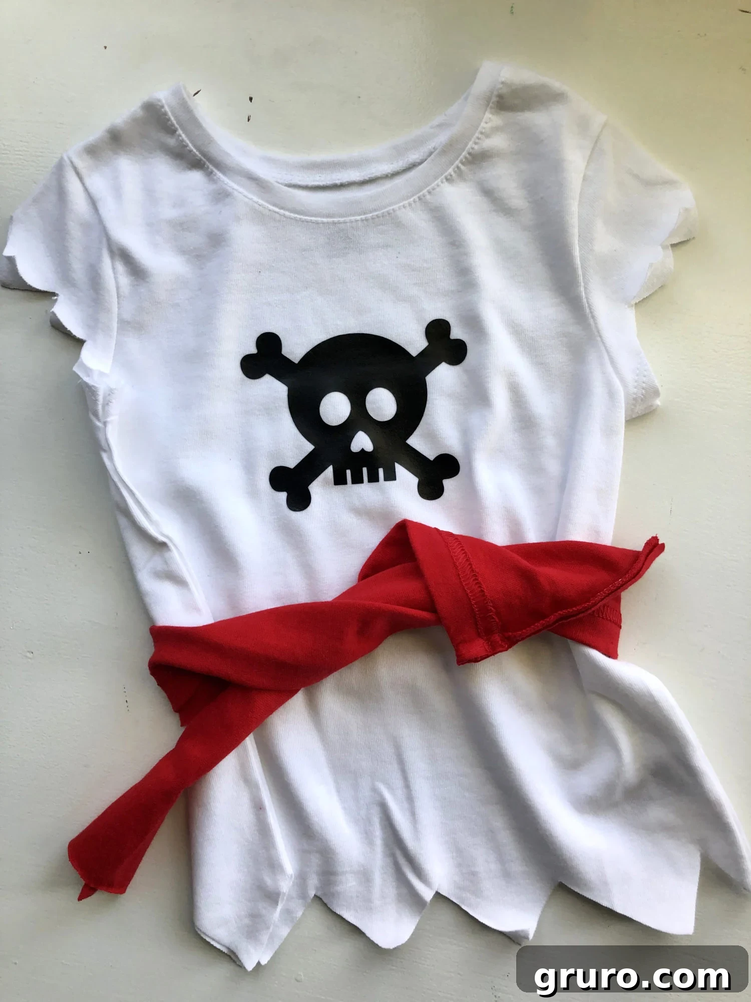 A white pirate shirt with a black skull design, accessorized with a red fabric belt, creating a simple yet effective pirate costume for a toddler.