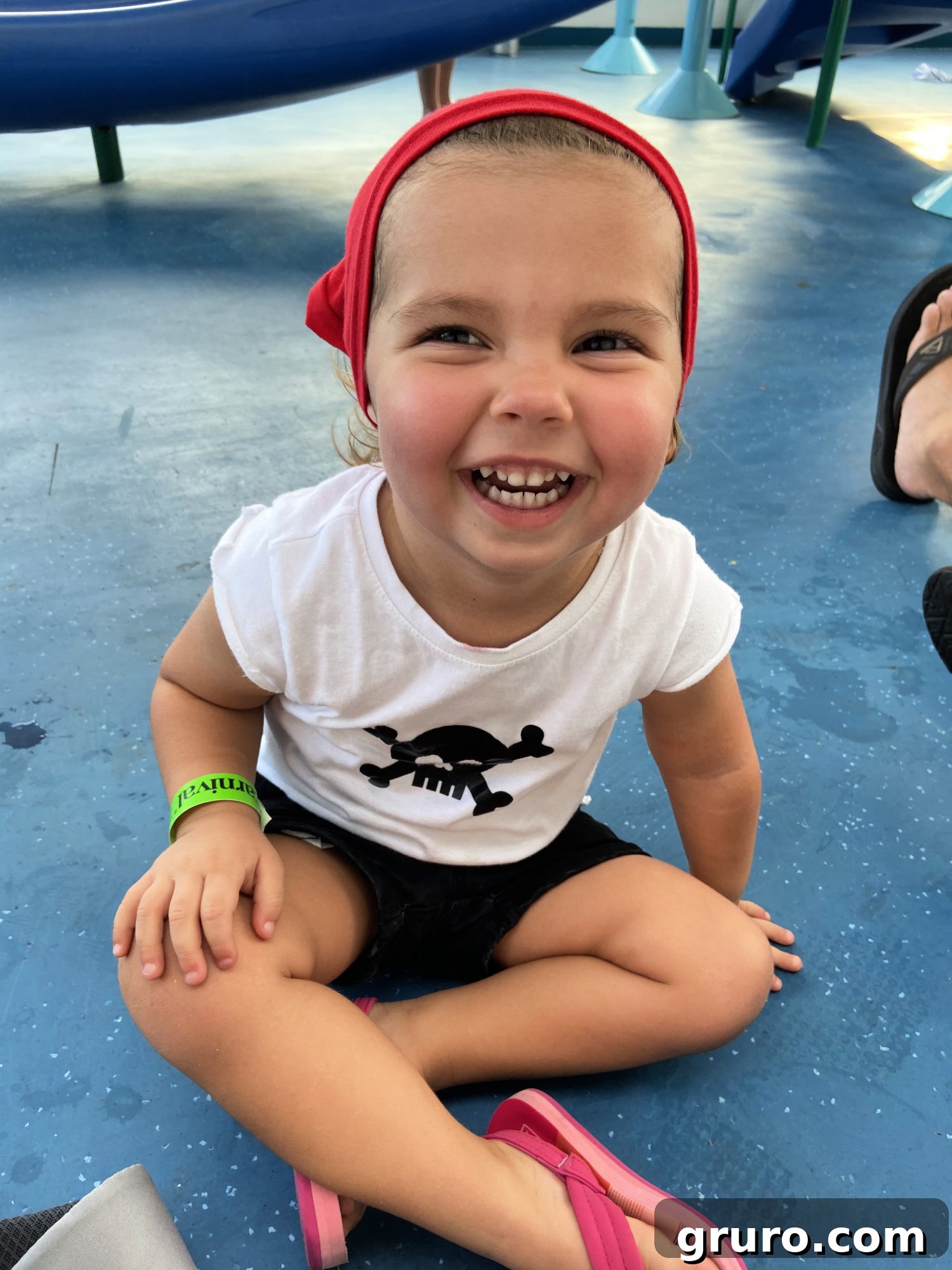 A toddler dressed in an adorable homemade pirate outfit, complete with a skull shirt, red vest, and du-rag, sitting on a speckled floor.