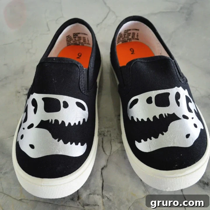Two black slip-on shoes adorned with custom dinosaur designs.