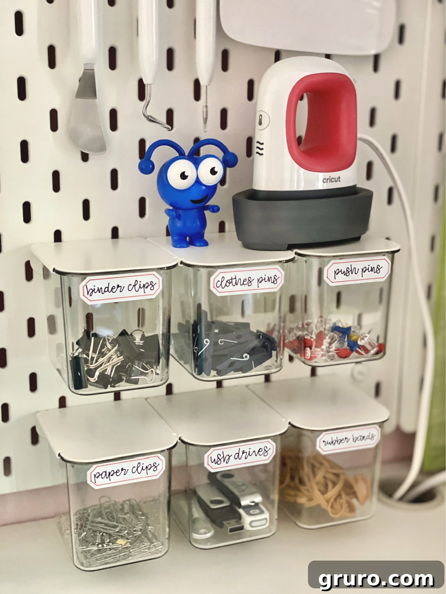 Clear bins neatly arranged on a pegboard, each adorned with elegant, hand-written style labels. A blue Cricut Joy machine and an EasyPress Mini are also visible, hinting at the tools used for this sophisticated organization system.