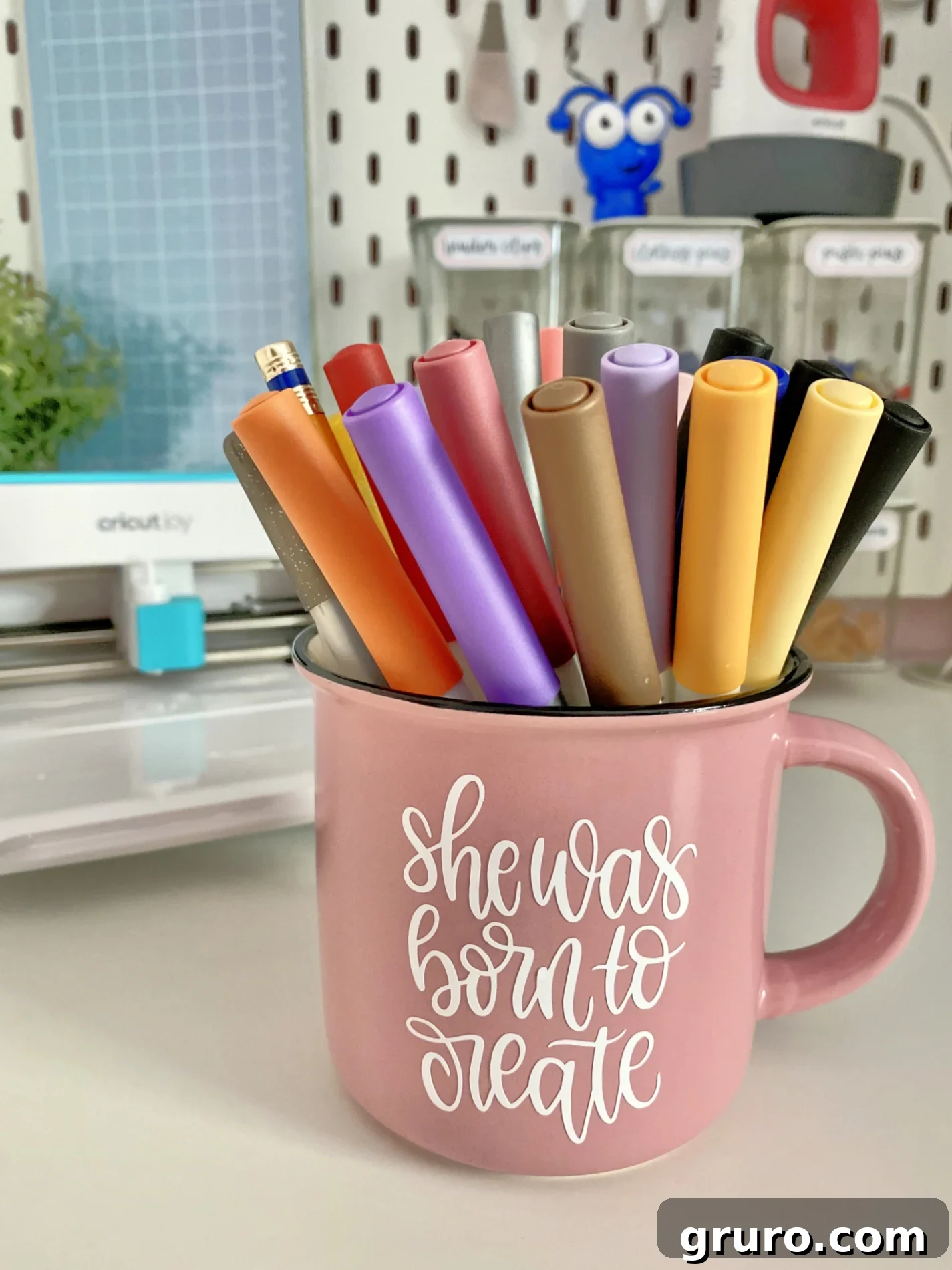 A vibrant pink mug proudly displaying the phrase "she was born to create" in white vinyl, filled with an assortment of pens. This inspiring mug serves as both a functional organizer and a creative desk accessory.