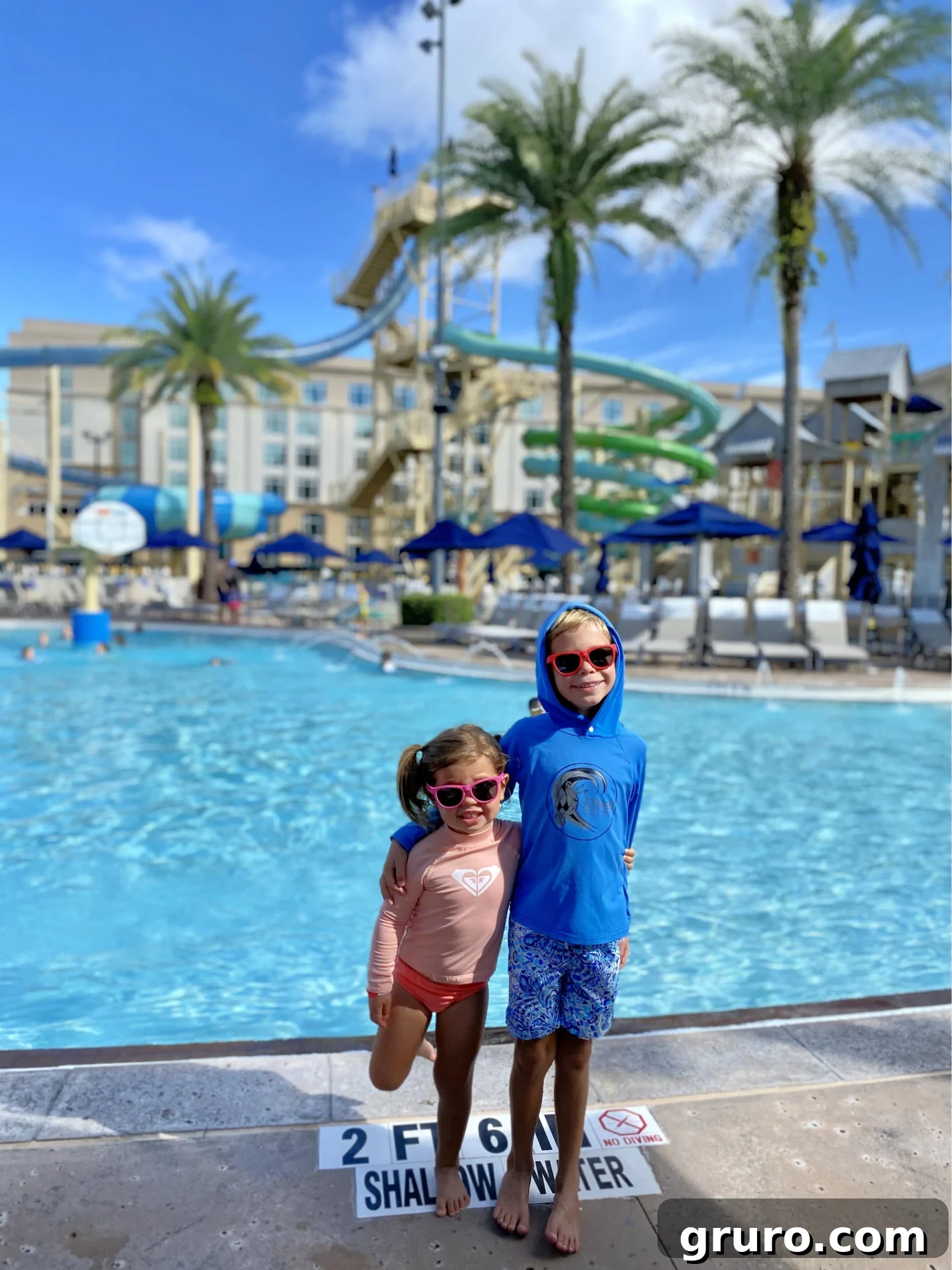 Two children in swimsuits and sunglasses standing in front of a swimming pool.