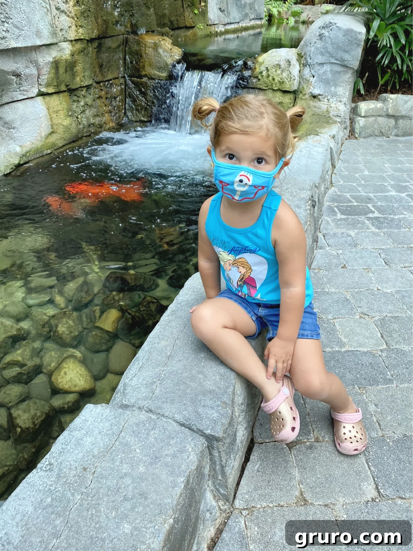 Little girl with blue mask on face sitting next to a koi pond. 
