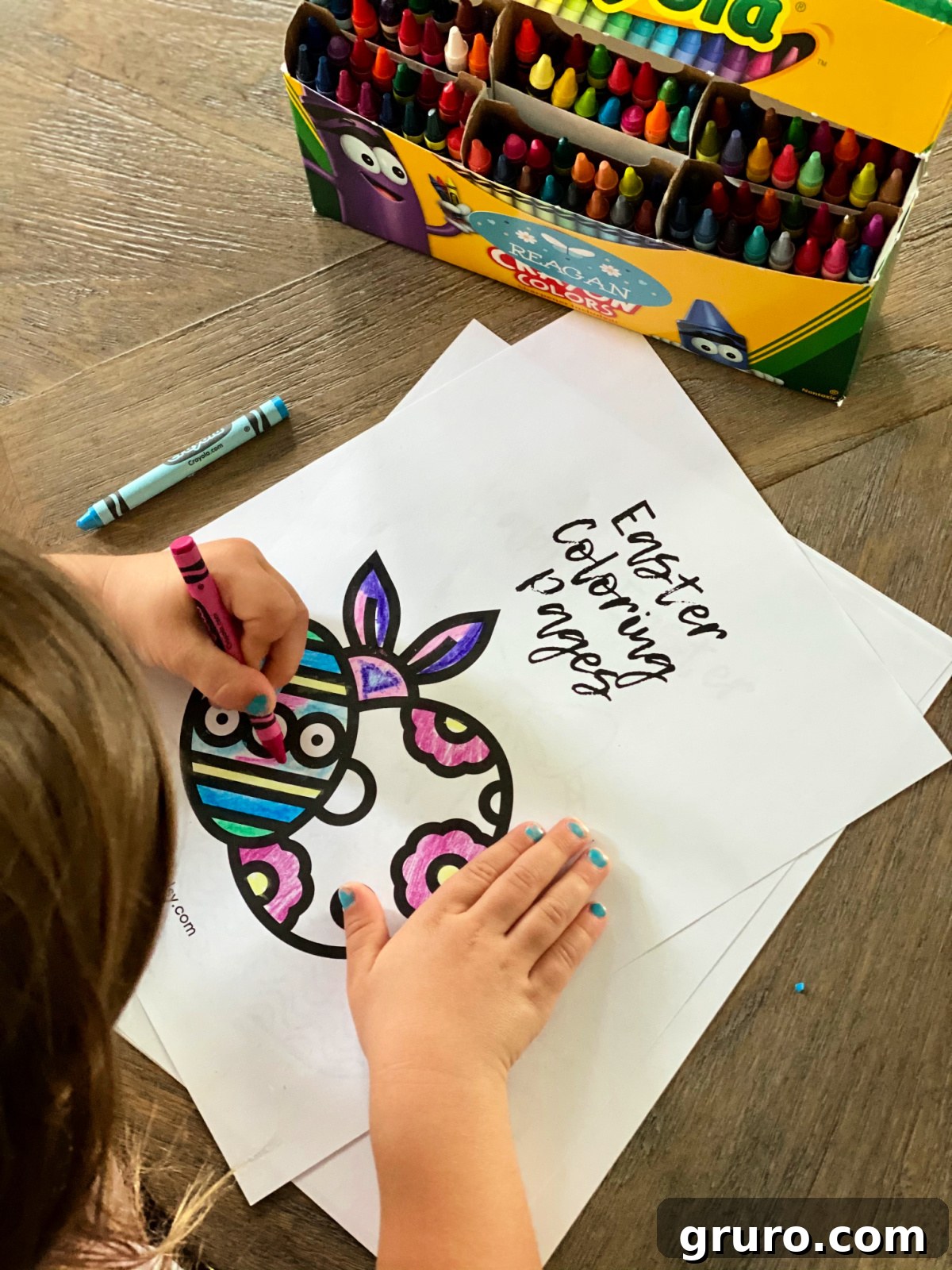 A little girl with blue fingernails intently coloring an Easter-themed page with colorful crayons, demonstrating focus and engagement.