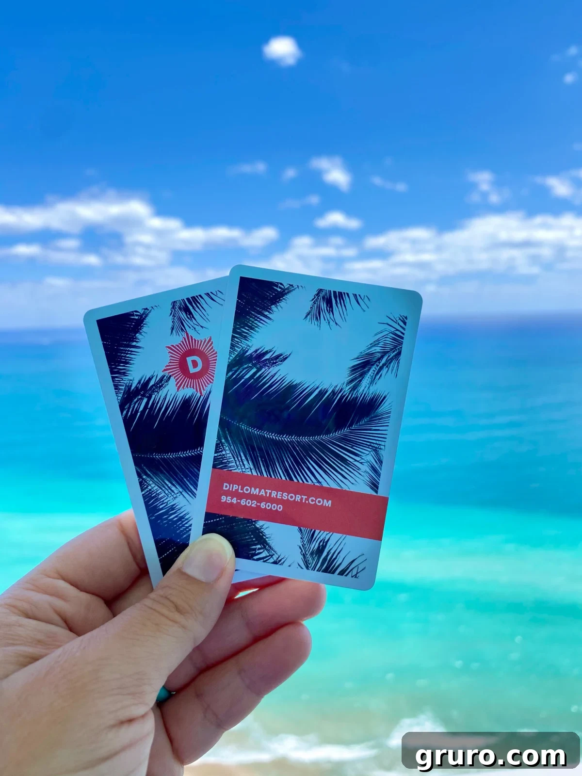 A female hand holding two elegant hotel room keys, with the glistening ocean visible in the background.