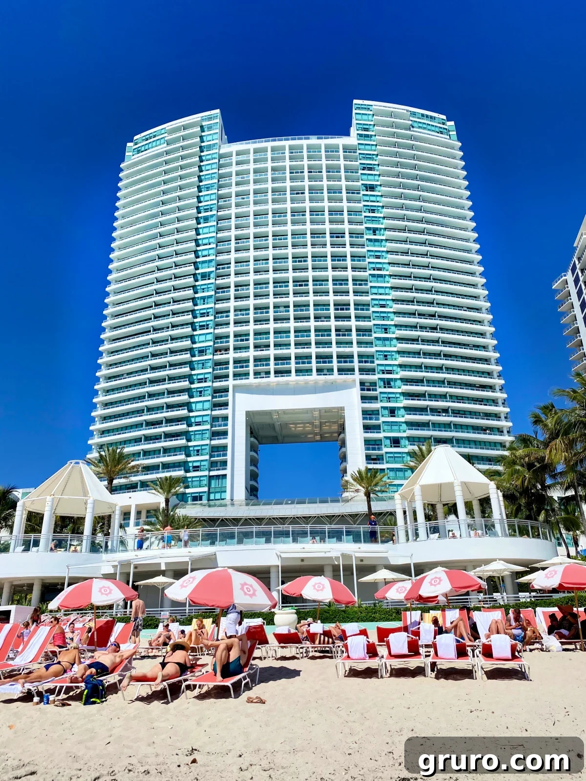 A large, modern hotel overlooking a vibrant beach scene with sunbathers enjoying lounge chairs and umbrellas.