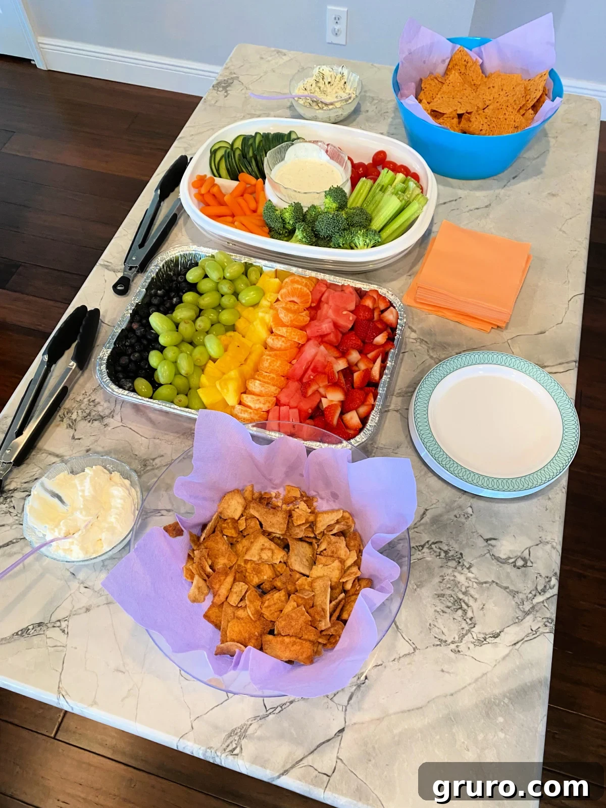 Rainbow fruit tray, veggie tray, chips and dip on marble kitchen island.
