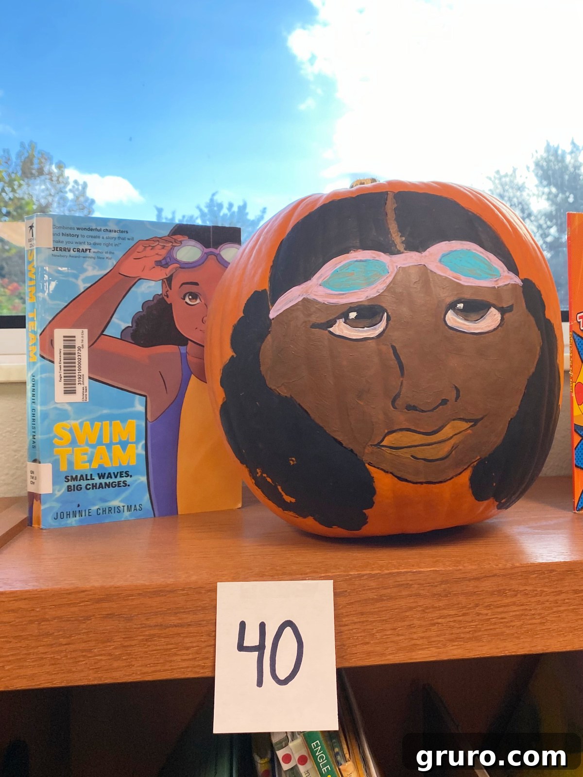 Orange pumpkin with an african american face on the pumpkin next to a book called "swim team"