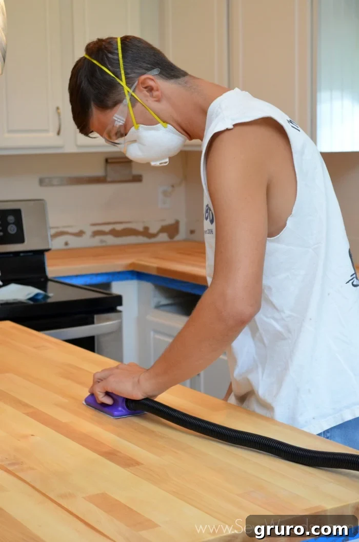 A man wearing a white cutoff shirt, dust mask, and safety goggles meticulously sanding butcher block countertops to achieve a smooth finish.