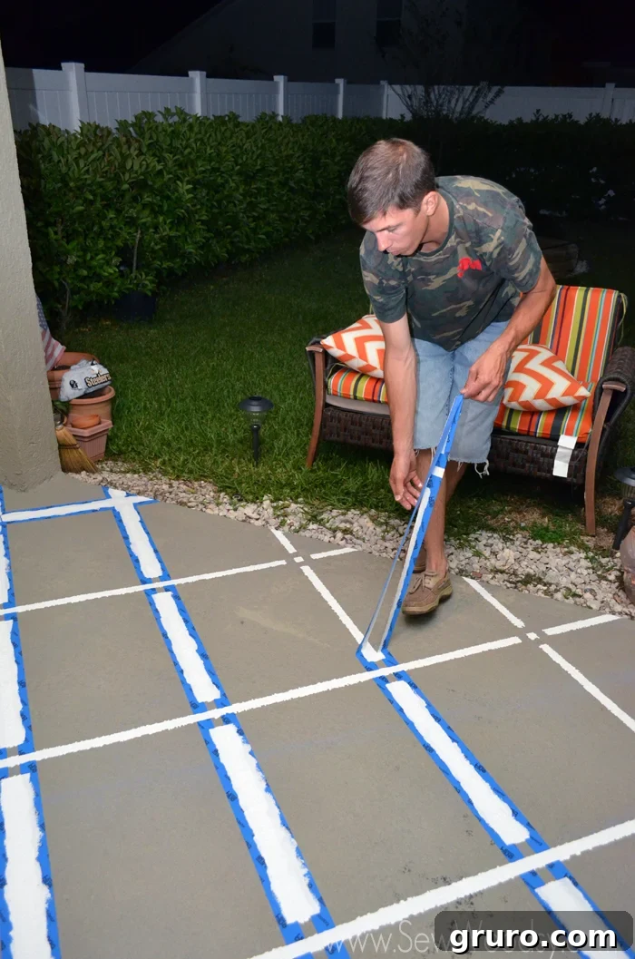 A person carefully pulling blue painter's tape off the concrete patio, revealing a crisp, newly painted geometric pattern underneath.