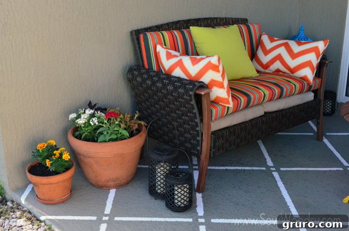 A beautifully decorated outdoor patio with a rattan couch, brightly colored cushions, potted flowers, lanterns, and a new white geometric pattern painted on the tan concrete floor.