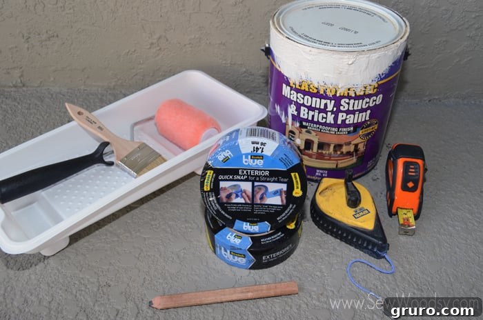 A collection of supplies neatly arranged on a patio, including a chalk reel, measuring tape, pencil, paint tray, exterior paint, and ScotchBlue painter's tape.