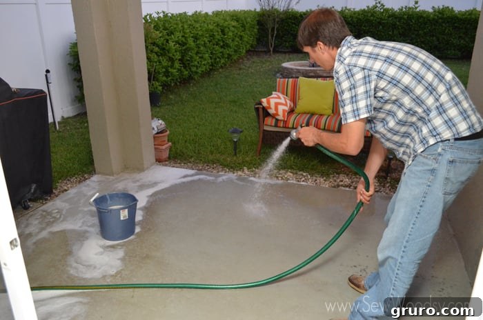 A man diligently spraying a concrete patio surface with a green hose, preparing it for painting.