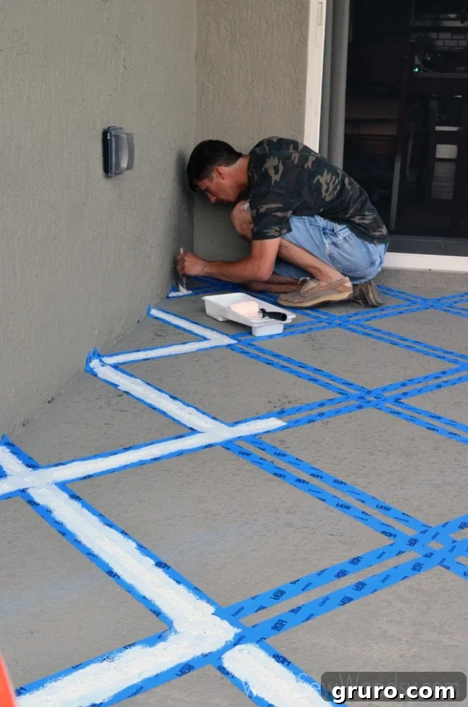 A person carefully using a paint brush to apply paint along the edges of blue painter's tape on a concrete patio.