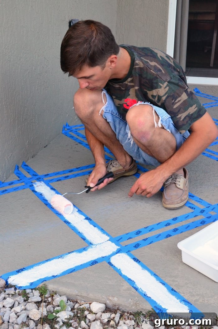 A person using a paint roller to apply paint evenly onto a taped-off section of a concrete patio.