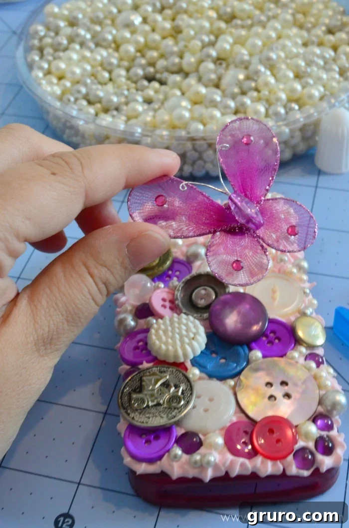 A Decoden phone case in progress, densely covered with an assortment of buttons, iridescent pearls, and sparkling purple gems, all embedded into the Strawberry Pink Mod Podge Collage Clay. This stage showcases the vibrant layering of various embellishments, creating a rich and intricate 3D texture across the entire surface of the case.