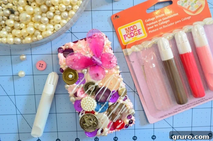 A close-up of a Decoden phone case with a diagonal drizzle of Collage Drizzle Paint applied over a section of the embedded buttons. The glossy drizzle adds a decorative, 'syrup-like' finish, enhancing the whimsical appeal of the meticulously arranged embellishments.