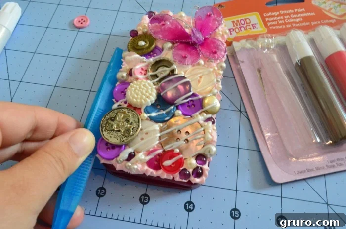 A stunning, completed Decoden phone case, densely adorned with an array of colorful buttons, shimmering pearls, and vibrant purple gems, all set in a Strawberry Pink Mod Podge Collage Clay base. A delicate 3D butterfly serves as a focal point, and glossy drizzle paint adds a final, intricate detail, making the phone case a unique piece of wearable art.
