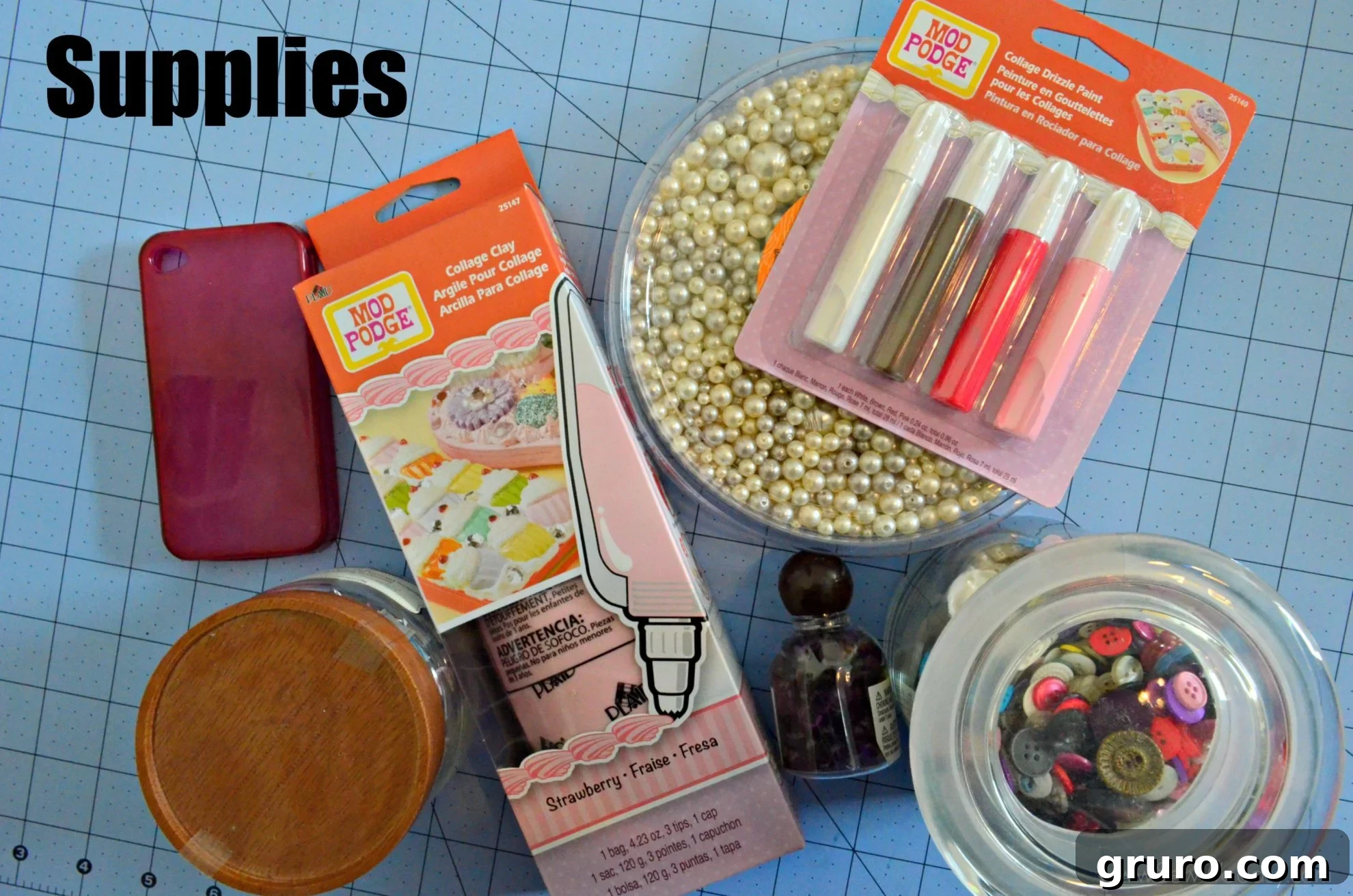 A flat lay photograph showcasing all the supplies needed for a Decoden phone case project. Items include a tube of Strawberry Pink Mod Podge Collage Clay with a star tip, a plain white cell phone case, a variety of colorful buttons, iridescent pearls, sparkling purple stones, a delicate 3D butterfly charm, and a bottle of Collage Drizzle Paint. These components are neatly arranged, indicating readiness for the crafting process.