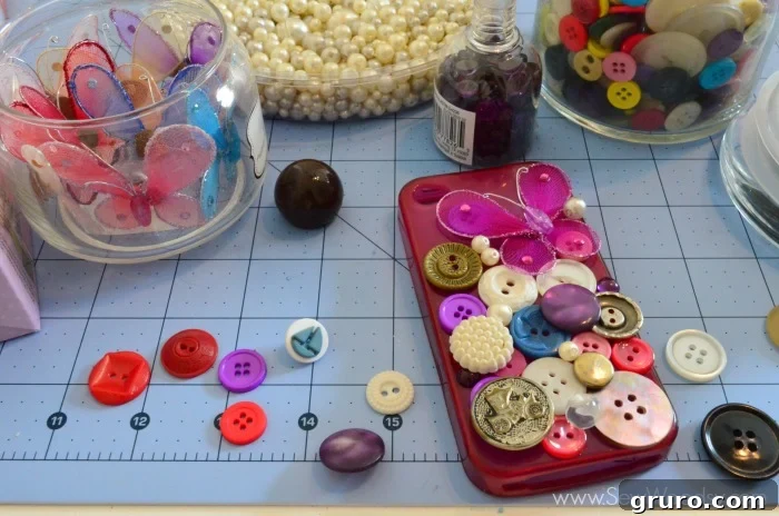 A plain cell phone case laid flat, with various decorative elements such as buttons, pearls, and purple stones carefully arranged on its surface. This preliminary step shows the crafter planning the design and layout of trinkets before applying the clay base, ensuring a balanced and aesthetically pleasing composition.