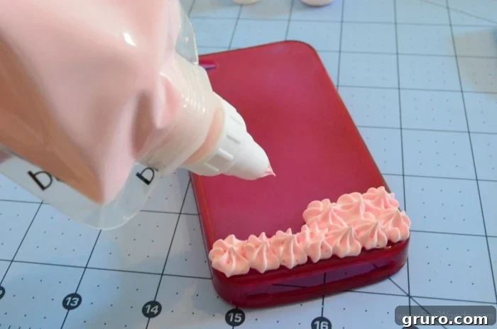 A hand expertly applying Strawberry Pink Mod Podge Collage Clay onto a cell phone case using a star tip, starting from the bottom edge. The image captures the meticulous process of creating the whipped cream base, showing the clay being piped in consistent, even swirls to cover the surface of the case.
