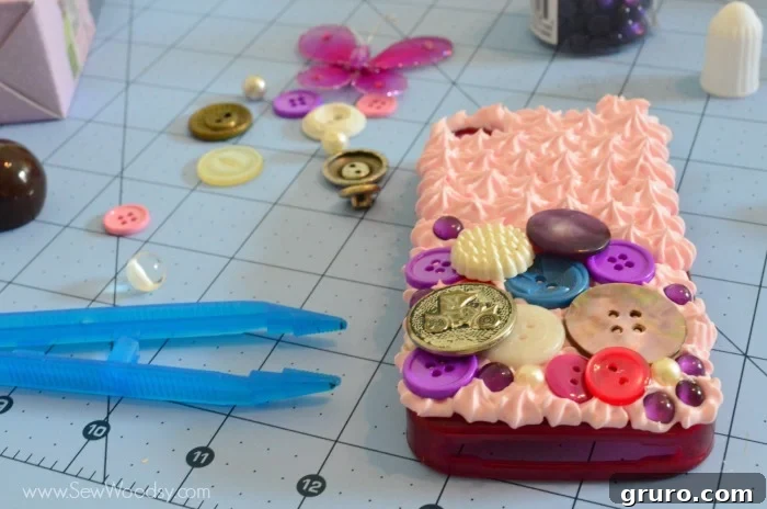 A close-up of a partially decorated Decoden phone case, with several colorful buttons already embedded into the wet Strawberry Pink Mod Podge Collage Clay. The image highlights the careful placement of the buttons, some assisted by craft tweezers for precision, as the crafter begins to build the 3D design.