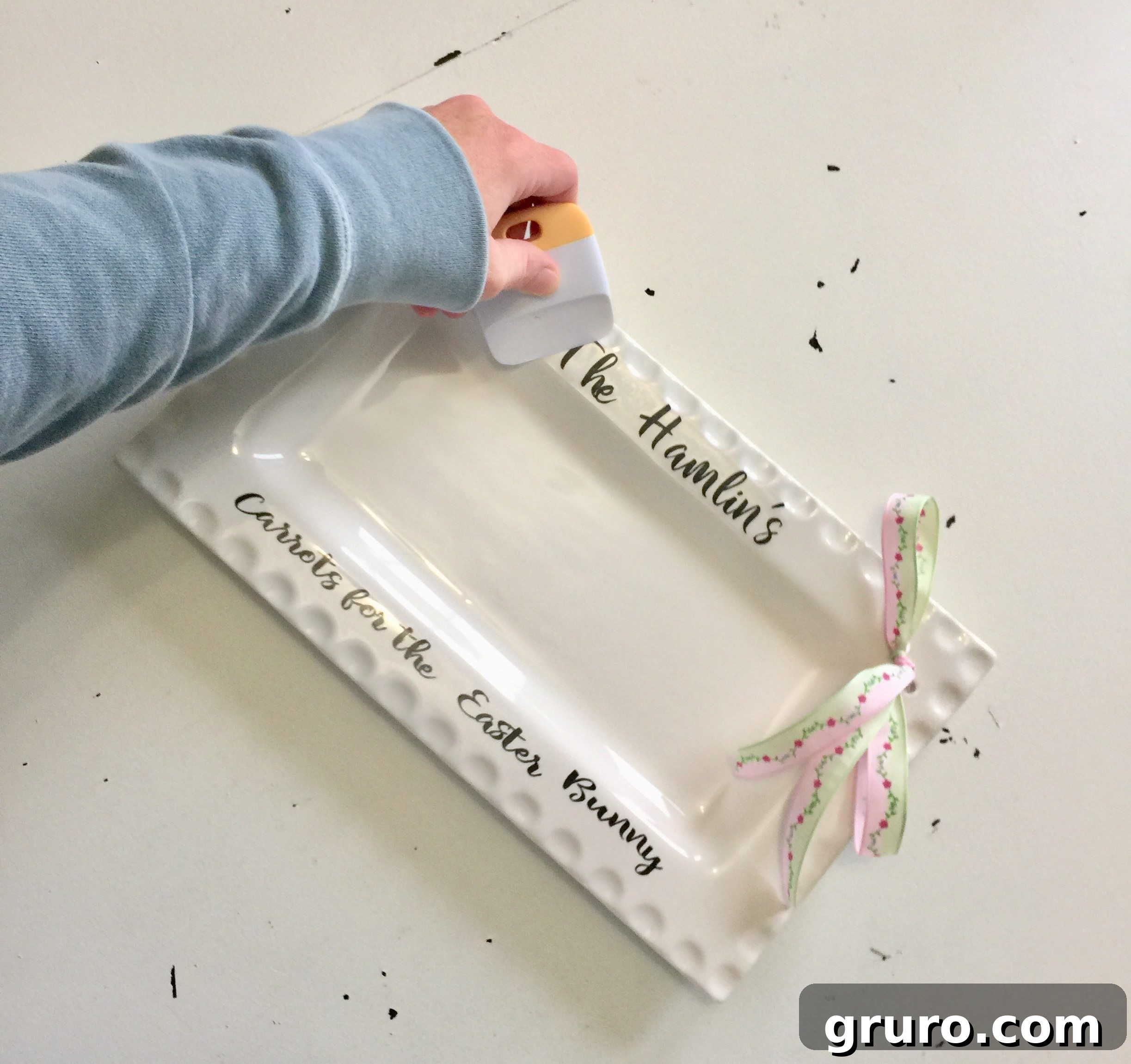 Hands carefully applying a vinyl design reading 'Carrots for the Easter Bunny' to a white ribbon platter using transfer tape and a scraper tool.